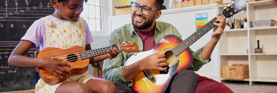 Happy male teacher assisting an african kid to play musical instrument in classroom at school. Happy teacher playing acoustic guitar and singing while having music class with girl. Cute little girl learn to play ukulele with male teacher.